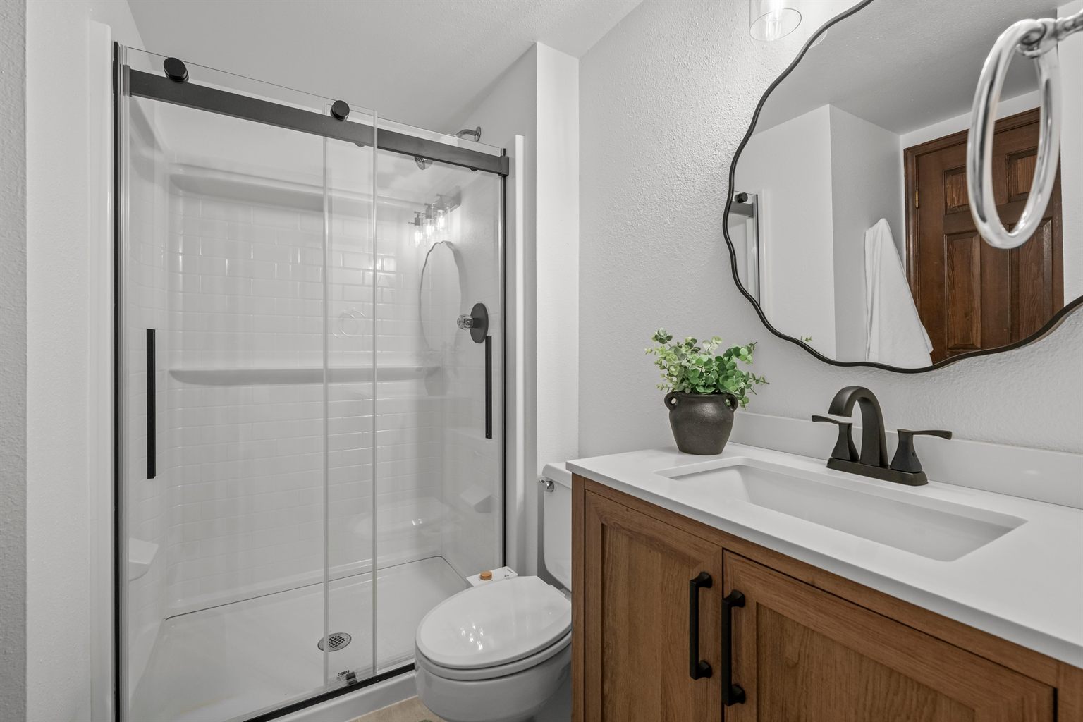 Main level bathroom after remodeling by SR Contractor LLC: white subway tile shower with black frame glass door, wood vanity and a black wavy mirror.